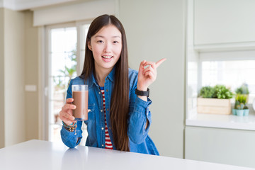 Beautiful Asian woman drinking a fresh glass of chocolate milkshake very happy pointing with hand and finger to the side