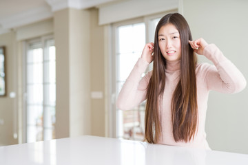Beautiful Asian woman wearing casual sweater on white table covering ears with fingers with annoyed expression for the noise of loud music. Deaf concept.