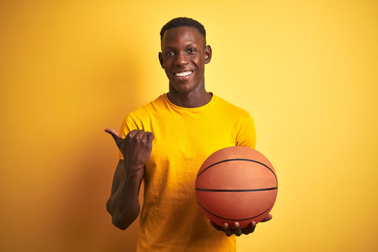 African american athlete man holding basketball ball standing over isolated yellow background pointing and showing with thumb up to the side with happy face smiling