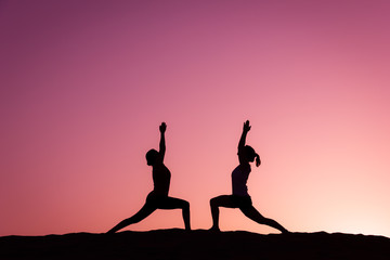 Yogi silhouettes on virabhadrasana on top of Maspalomas sand dunes at colorful sunset. Couple of women practice warrior pose at pink twilight in Canary Islands, Spain