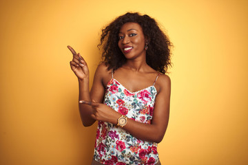 African american woman wearing floral summer t-shirt over isolated yellow background smiling and looking at the camera pointing with two hands and fingers to the side.