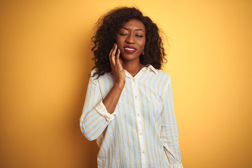 African american woman wearing striped shirt standing over isolated yellow background touching mouth with hand with painful expression because of toothache or dental illness on teeth. Dentist concept.