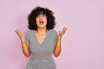 Young arab woman with curly hair wearing striped dress over isolated pink background crazy and mad shouting and yelling with aggressive expression and arms raised. Frustration concept.