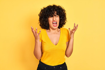 Young arab woman with curly hair wearing t-shirt standing over isolated yellow background celebrating mad and crazy for success with arms raised and closed eyes screaming excited. Winner concept