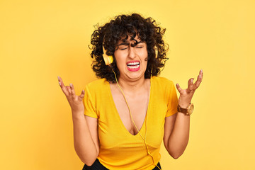 Arab woman with curly hair listening to music using headphones over isolated yellow background celebrating mad and crazy for success with arms raised and closed eyes screaming excited. Winner concept