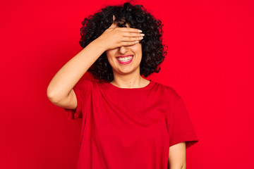 Fototapeta na wymiar Young arab woman with curly hair wearing casual t-shirt over isolated red background smiling and laughing with hand on face covering eyes for surprise. Blind concept.
