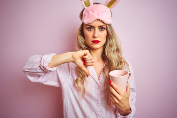 Young woman wearing pajama and mask drinking a cup of coffee over pink isolated background with angry face, negative sign showing dislike with thumbs down, rejection concept