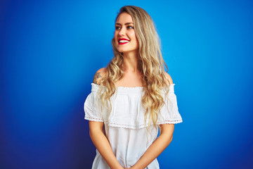 Young beautiful woman wearing white dress standing over blue isolated background looking away to side with smile on face, natural expression. Laughing confident.