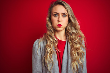 Young beautiful business woman wearing elegant jacket standing over red isolated background puffing cheeks with funny face. Mouth inflated with air, crazy expression.