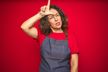 Middle age senior woman wearing apron uniform over red isolated background making fun of people...