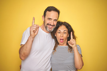 Beautiful middle age couple together standing over isolated yellow background surprised with an idea or question pointing finger with happy face, number one