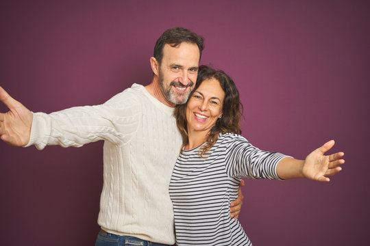 Beautiful Middle Age Couple Wearing Winter Sweater Over Isolated Purple Background Looking At The Camera Smiling With Open Arms For Hug. Cheerful Expression Embracing Happiness.