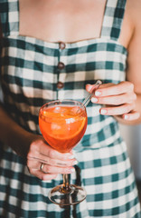 Young woman in checkered dress holding glass of Aperol Spritz aperitif drink cocktail with orange...