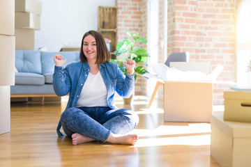 Young plus size woman sitting on the floor around cardboard boxes moving to a new home looking...