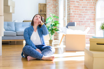 Young plus size woman sitting on the floor around cardboard boxes moving to a new home with hand on chin thinking about question, pensive expression. Smiling with thoughtful face. Doubt concept.