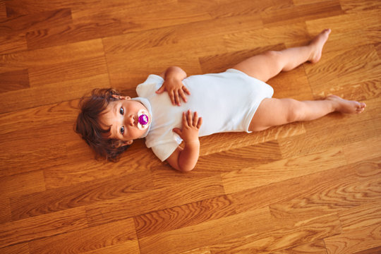 Beautiful toddler child girl wearing white bodysuit lying down on the floor using pacifier