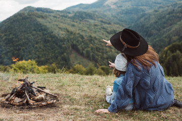 Fototapeta na wymiar Beautiful young mother playing with daughter near to campfire in mountains in wild national park forest. Traveler family relaxing after mountaineering. Caucasian female with baby at mountains
