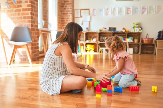 Beautiful teacher and blond toddler girl building tower using plastic blocks at kindergarten