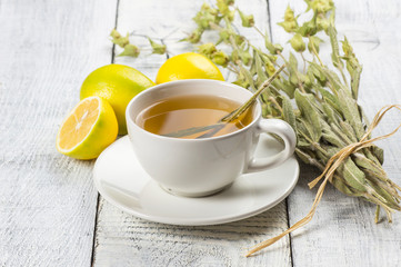 White cup of herbal sage tea with dried sage leaves and lemon on white wooden rustic background. Herbal tea hot drink concept,  Salvia officinalis
