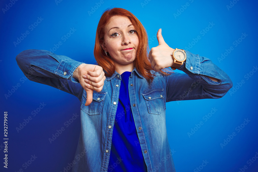 Sticker Young beautiful redhead woman wearing denim shirt standing over blue isolated background Doing thumbs up and down, disagreement and agreement expression. Crazy conflict