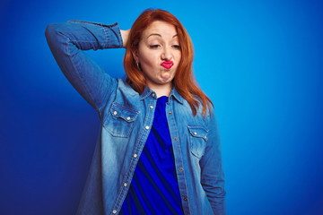 Young beautiful redhead woman wearing denim shirt standing over blue isolated background confuse...