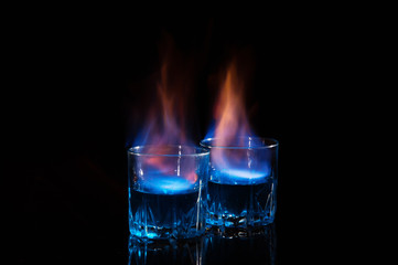 Close up, macro. Two glass glasses with blue alcohol with a flame of burning fire. Black...