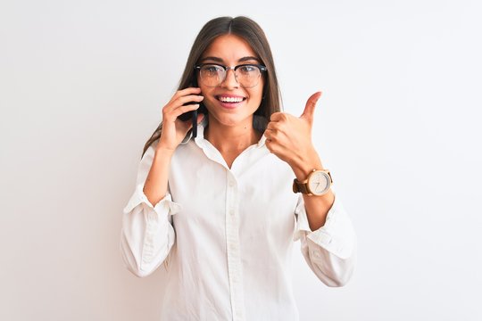 Young businesswoman wearing glasses talking on smartphone over isolated white background happy with big smile doing ok sign, thumb up with fingers, excellent sign