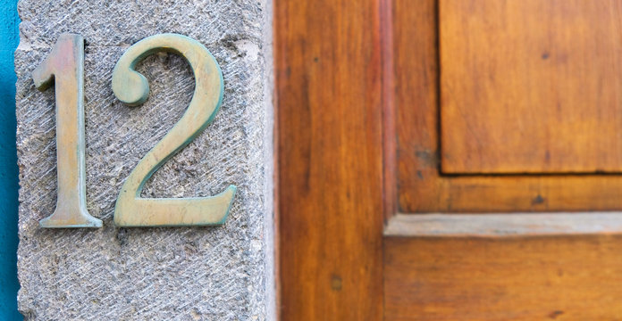 Number 12 / twelve, elegant metal digits house number on the wall, warm wood door detail in soft background.