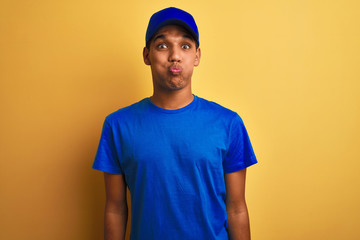 Young handsome arab delivery man standing over isolated yellow background puffing cheeks with funny face. Mouth inflated with air, crazy expression.