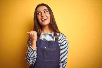 Young beautiful baker woman wearing apron standing over isolated yellow background smiling with happy face looking and pointing to the side with thumb up.