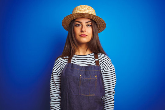 Young beautiful baker woman wearing apron and hat standing over isolated blue background with serious expression on face. Simple and natural looking at the camera.