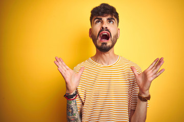 Young man with tattoo wearing striped t-shirt standing over isolated yellow background crazy and...