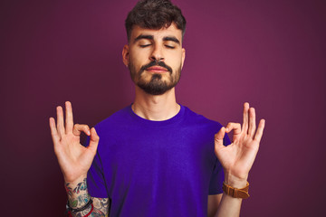 Young man with tattoo wearing t-shirt standing over isolated purple background relax and smiling with eyes closed doing meditation gesture with fingers. Yoga concept.