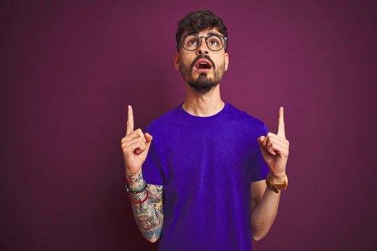 Young man with tattoo wearing t-shirt and glasses standing over isolated purple background amazed and surprised looking up and pointing with fingers and raised arms.