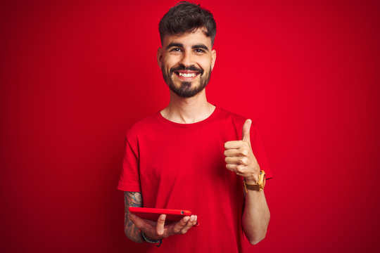Young man with tattoo using tablet standing over isolated red background happy with big smile doing ok sign, thumb up with fingers, excellent sign