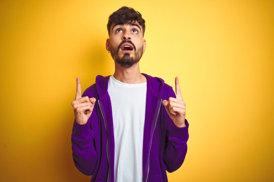 Young man with tattoo wearing sport purple sweatshirt over isolated yellow background amazed and surprised looking up and pointing with fingers and raised arms.