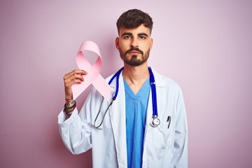 Young doctor man with tattoo holding cancer ribbon standing over isolated pink background with a confident expression on smart face thinking serious