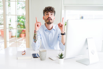 Young business man working using computer amazed and surprised looking up and pointing with fingers and raised arms.