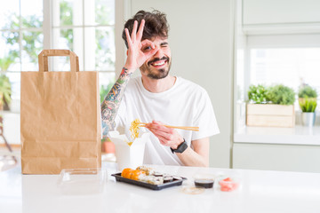 Young man eating asian sushi from home delivery with happy face smiling doing ok sign with hand on eye looking through fingers