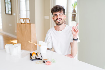 Young man eating asian sushi from home delivery with a big smile on face, pointing with hand and finger to the side looking at the camera.