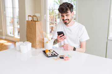 Young man eating asian sushi from home delivery and ordering food using smartphone app with a happy face standing and smiling with a confident smile showing teeth