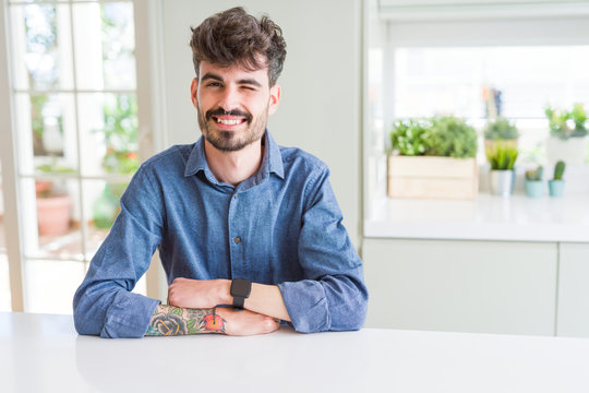 Young man wearing casual shirt sitting on white table winking looking at the camera with sexy expression, cheerful and happy face.