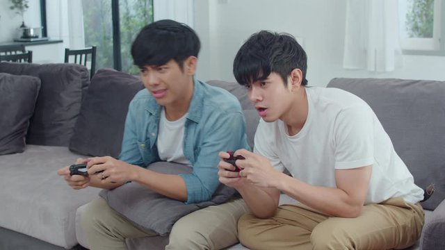 Young Asian gay couple play games at home, Teen korean LGBTQ men using joystick having funny happy moment together on sofa in living room at house. Slow motion shot.