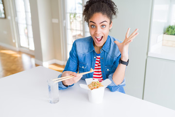 Young african american woman eating asian noodles from delivery box very happy and excited, winner expression celebrating victory screaming with big smile and raised hands