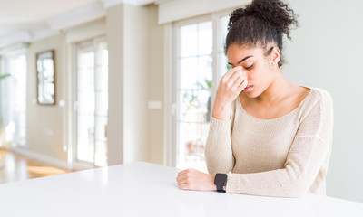 Beautiful young african american woman with afro hair sitting on table at home tired rubbing nose and eyes feeling fatigue and headache. Stress and frustration concept.