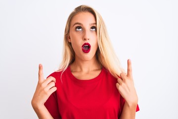Young beautiful woman wearing red casual t-shirt standing over isolated white background amazed and surprised looking up and pointing with fingers and raised arms.
