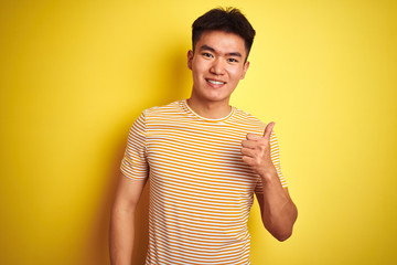 Young asian chinese man wearing t-shirt standing over isolated yellow background doing happy thumbs up gesture with hand. Approving expression looking at the camera showing success.