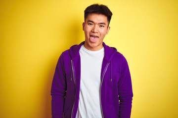Young asian chinese man wearing purple sweatshirt standing over isolated yellow background sticking tongue out happy with funny expression. Emotion concept.