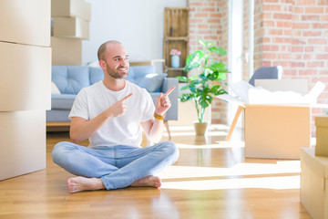 Young bald man sitting on the floor around cardboard boxes moving to a new home smiling and looking...