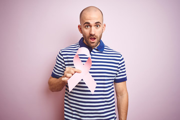 Young man holding pink brest cancer ribbon over isolated background scared in shock with a surprise...